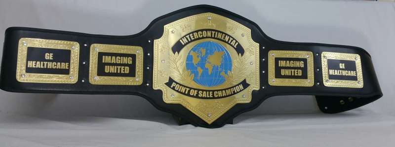 A novelty black and gold championship belt with five plates, featuring humorous text such as 'GE HEALTHCARE', 'IMAGING UNITED', and 'POINT OF SALE CHAMPION' on a light gray background.