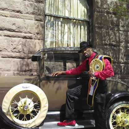 A man in a black hat, red jacket, and black pants sits on a vintage car, holding a championship belt. The car has cream-colored tires with gold-spoked rims.
